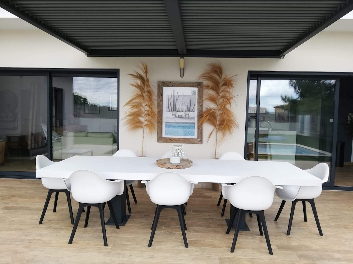 An outdoor dining area is presented under a covered terrace, featuring a long white table surrounded by eight modern white chairs. Large glass doors provide a view of the surrounding space, with decorative elements and natural textures enhancing the setting.