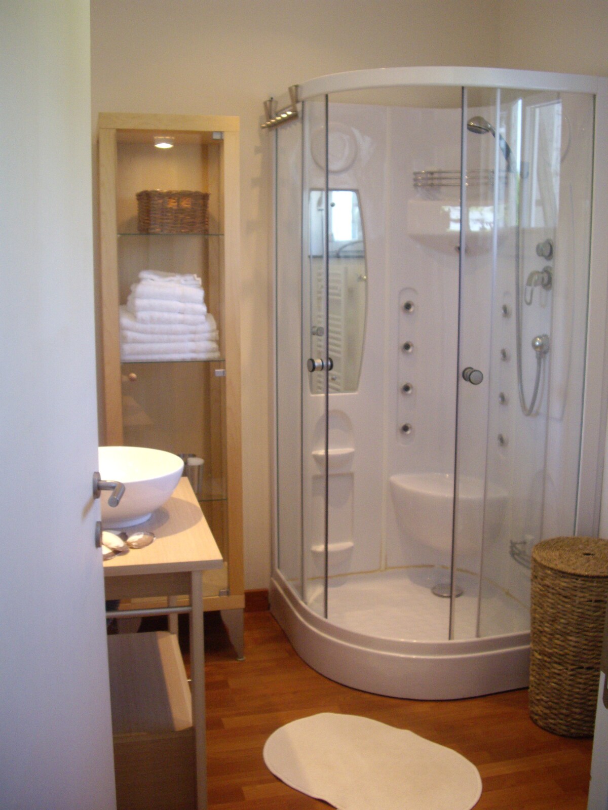 A compact bathroom features a glass-enclosed shower with multiple body jets and a handheld showerhead. A modern sink is situated atop a light wood vanity, with neatly stacked white towels displayed on an adjacent shelf. A decorative basket adds texture to the space.