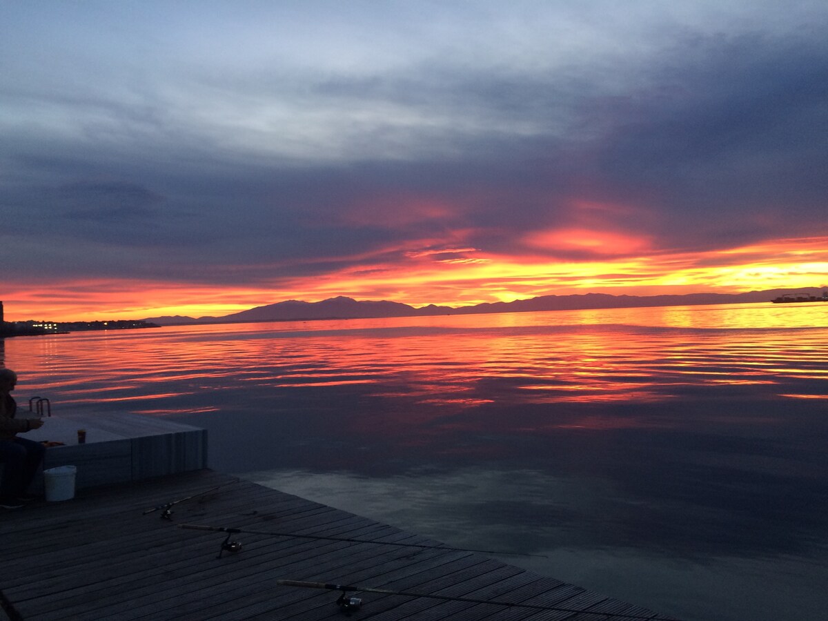 A serene waterfront scene is depicted, featuring a calm body of water reflecting vibrant hues of orange, pink, and purple from the sunset. Silhouettes of distant mountains are visible on the horizon, while a wooden dock is presented in the foreground.
