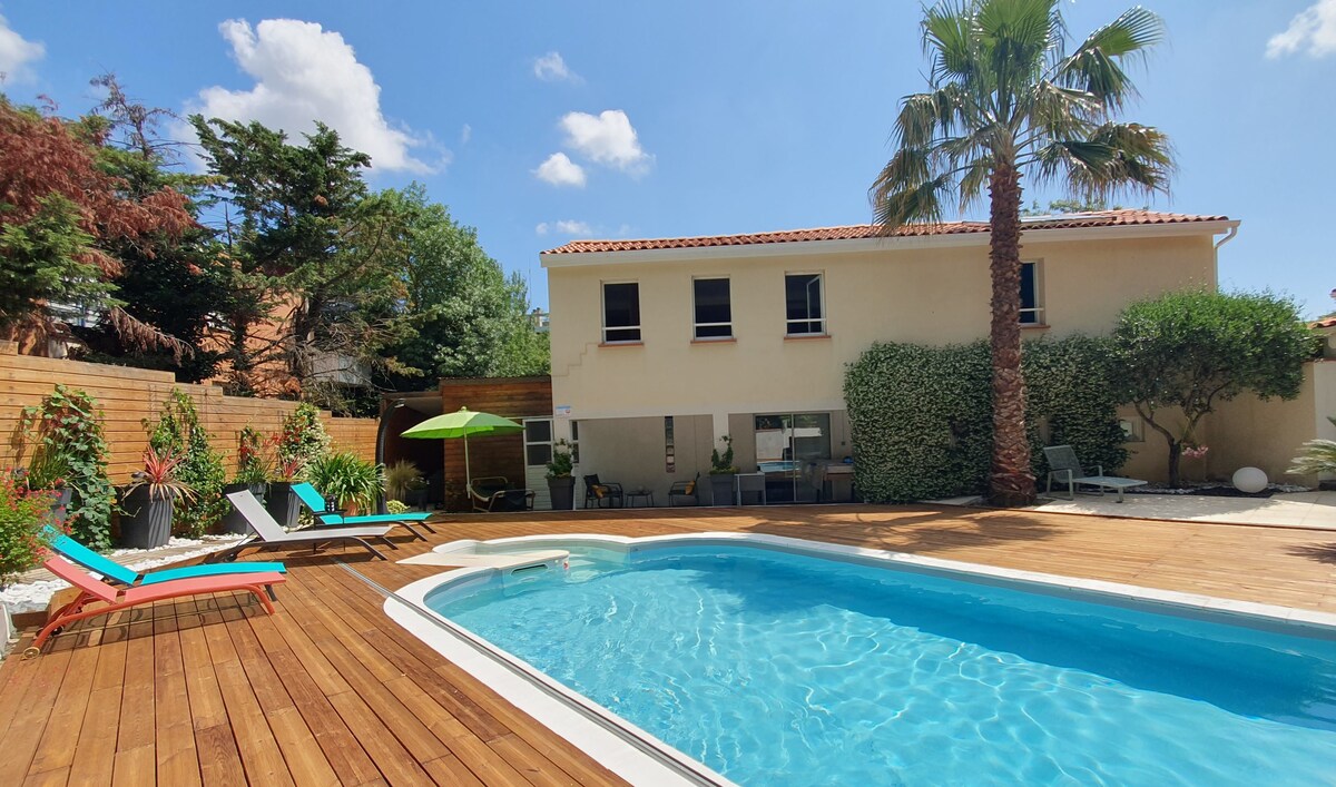 A private outdoor area features a rectangular swimming pool surrounded by wooden decking. Colorful lounge chairs are positioned nearby, with a palm tree providing shade. The house, with large windows, is set against a backdrop of greenery and decorative plants.