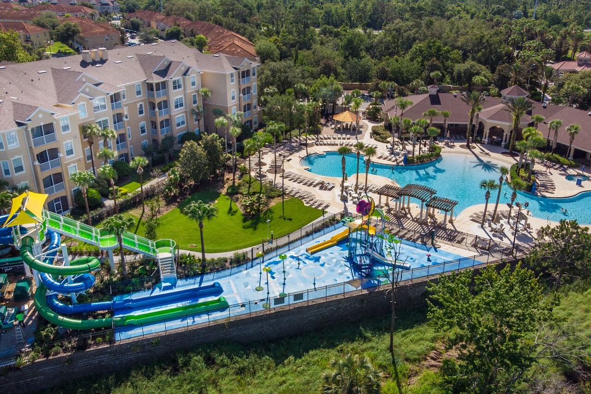 An aerial view of a resort features an expansive water park with multiple slides in vibrant colors. A large pool with lounge chairs surrounds the area, complemented by lush landscaping and palm trees. Nearby, shaded cabanas provide additional relaxation space.
