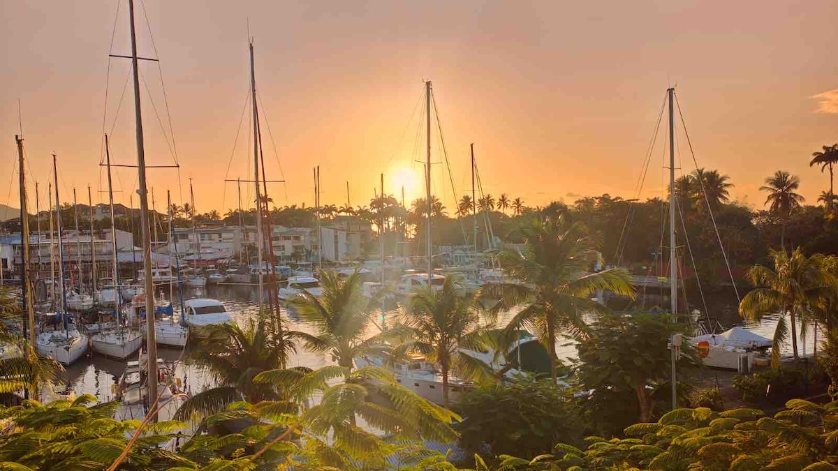 A tranquil marina scene is presented at sunset, with sailboats anchored gently on the water. Silhouetted palm trees frame the image, while warm hues of orange and pink reflect off the surface. The overall atmosphere conveys relaxation and natural beauty.