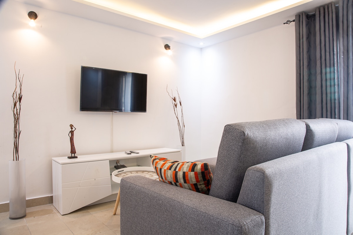 A modern living area is highlighted by a spacious gray sofa positioned towards a mounted flat-screen TV. A circular coffee table rests in front, complemented by a decorative accent pillow. Contemporary wall decorations and soft lighting create a clean and inviting ambiance.
