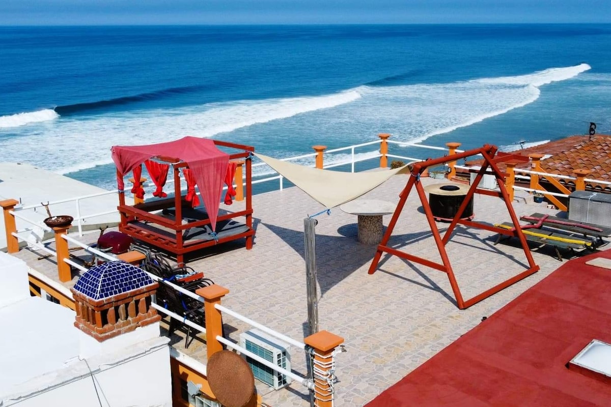 The rooftop deck provides a view of the ocean waves, featuring a shaded seating area adorned with draped fabric. A wooden swing set is positioned nearby, while sun loungers are arranged for relaxation. The tiled surface complements the coastal ambiance.