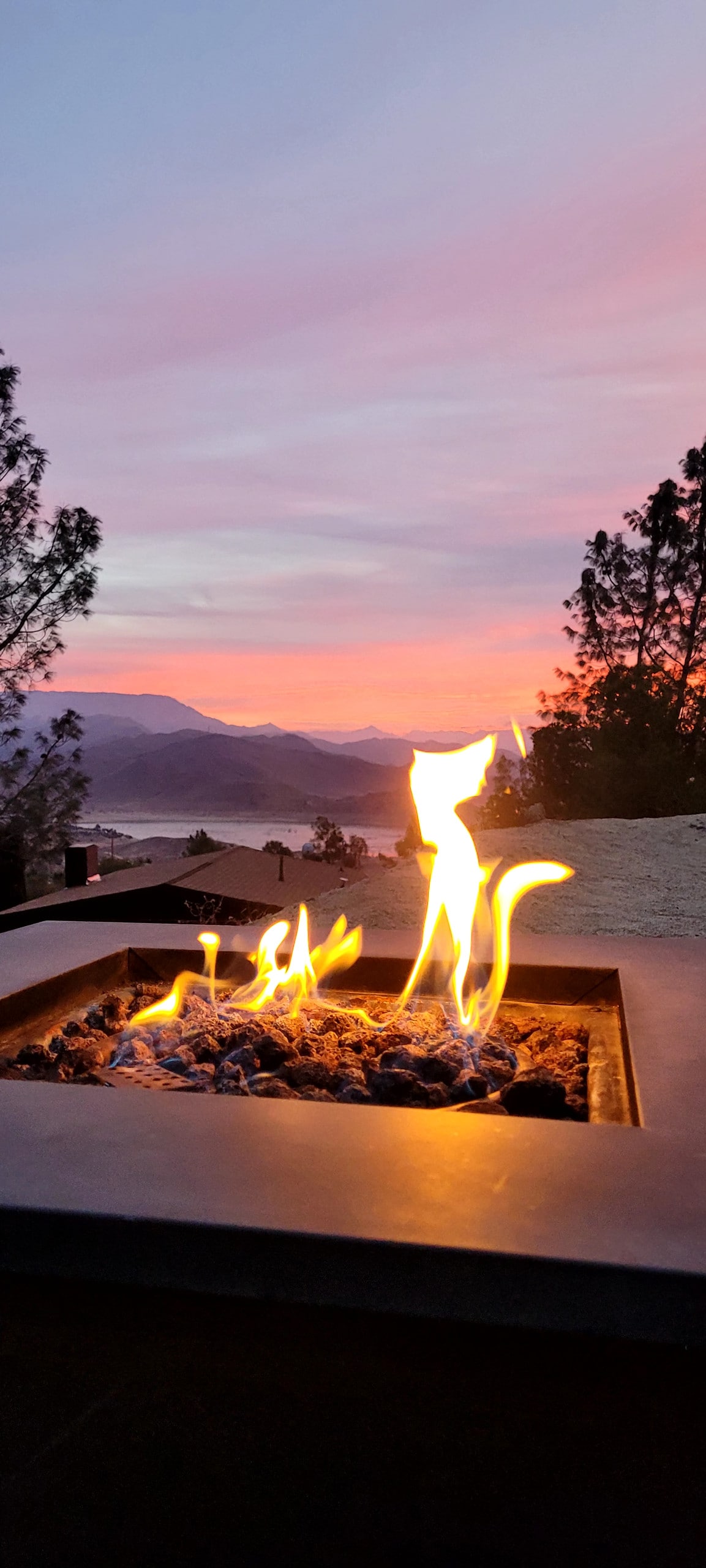 A propane fire pit is positioned on a deck, with flames gently flickering above the stone base. The background features a serene sunset over mountains, encompassing a gradient of pink and orange hues against a soft purple sky, enhancing the tranquil outdoor setting.