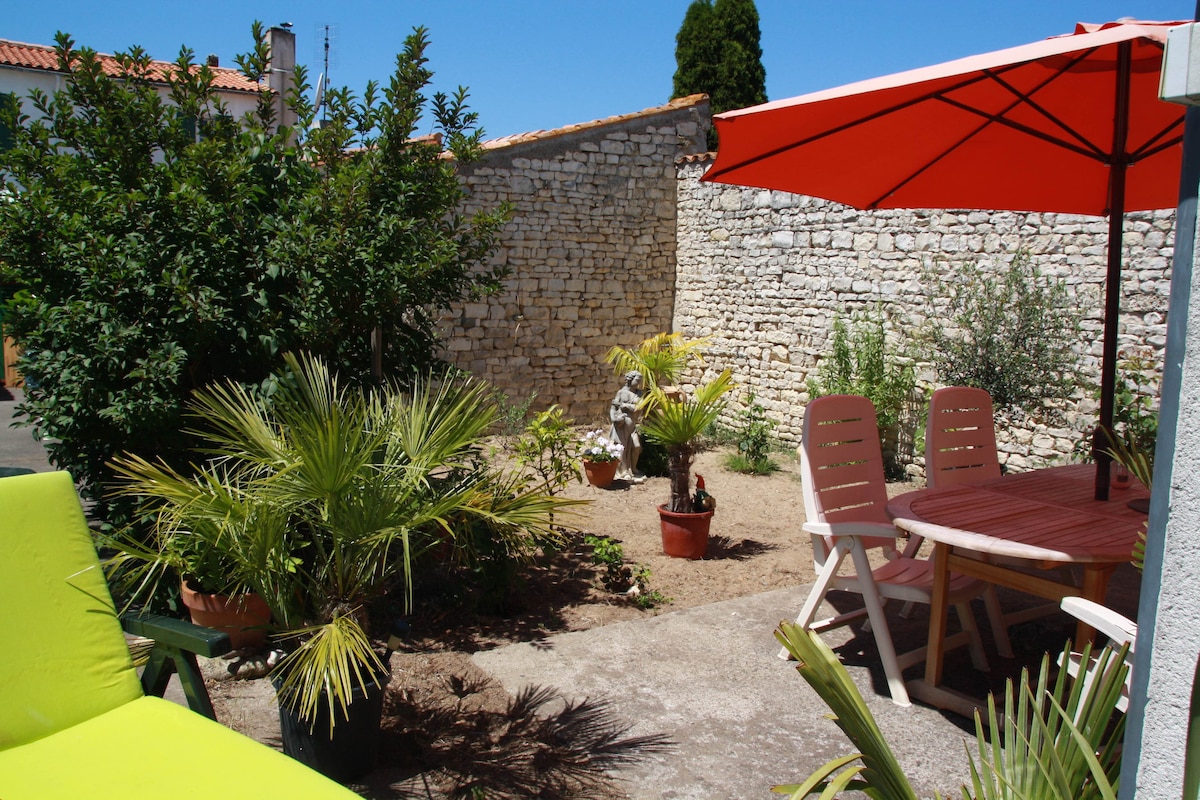 A garden space is visible, featuring a wooden dining table under a large red umbrella. Potted plants, including palms, are arranged around the area. A stone wall provides privacy, and a seating area with green lounge chairs adds comfort.