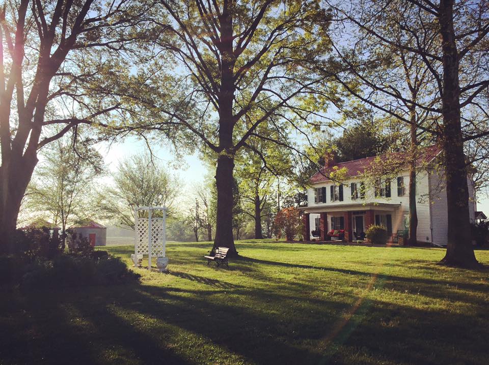A historic antebellum house is set on sprawling green grounds, framed by mature trees. Natural light filters through the branches, casting a gentle glow on the lush lawn. A white trellis stands in the foreground, hinting at the property's charm. Seating is arranged on the porch.