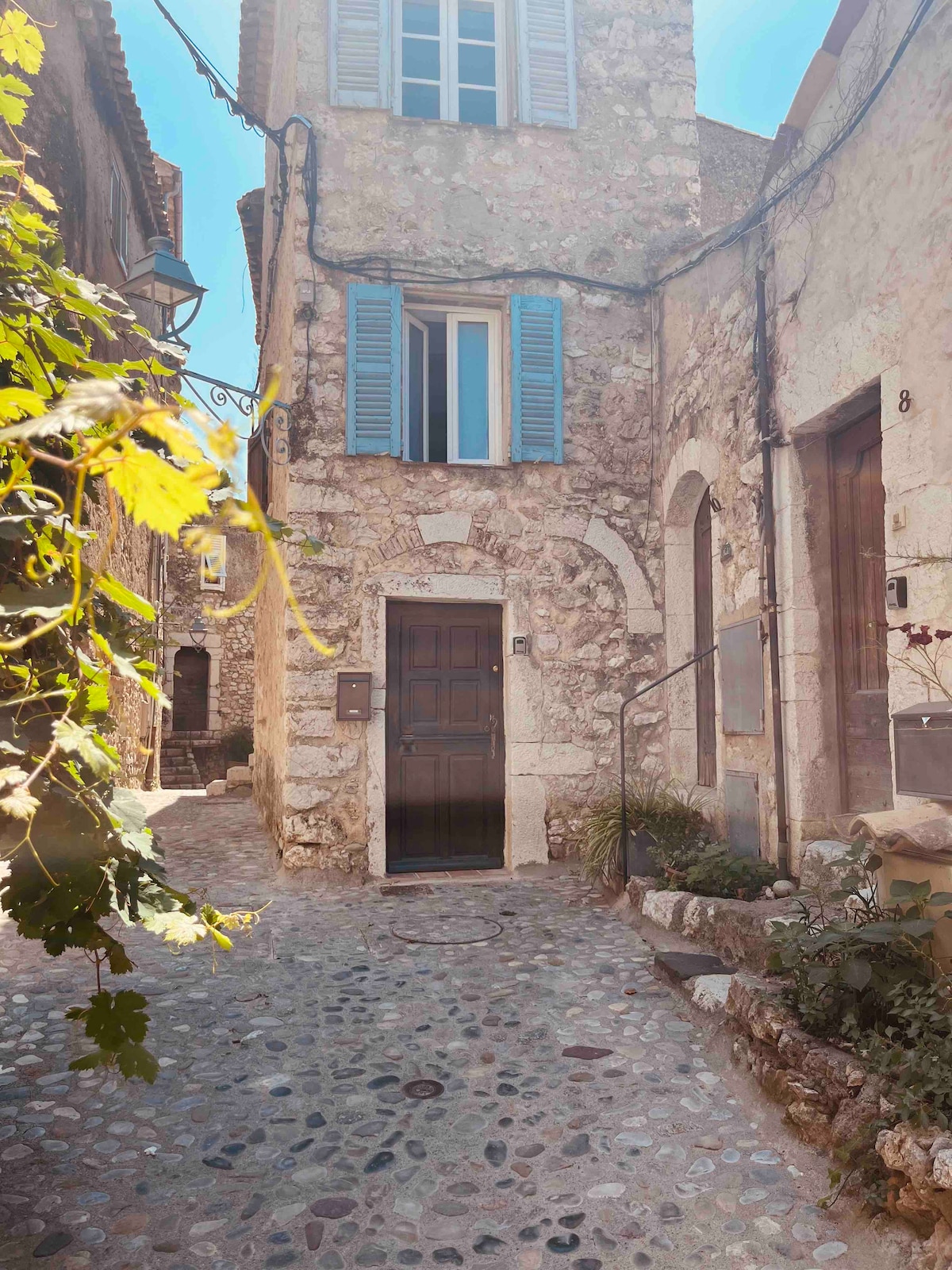 The image shows a charming stone building with light blue shutters, situated on a cobblestone street. A wooden door adorned with a decorative archway serves as the main entrance. Green foliage is visible in the foreground, enhancing the rustic appeal of the setting.