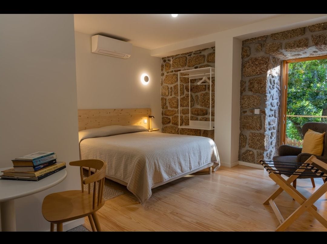 A spacious sleeping area features a large bed with a light-colored coverlet, complemented by a stone accent wall. A wooden chair and a folding chair provide seating options, while natural light flows through the open doorway that leads to an outdoor view.