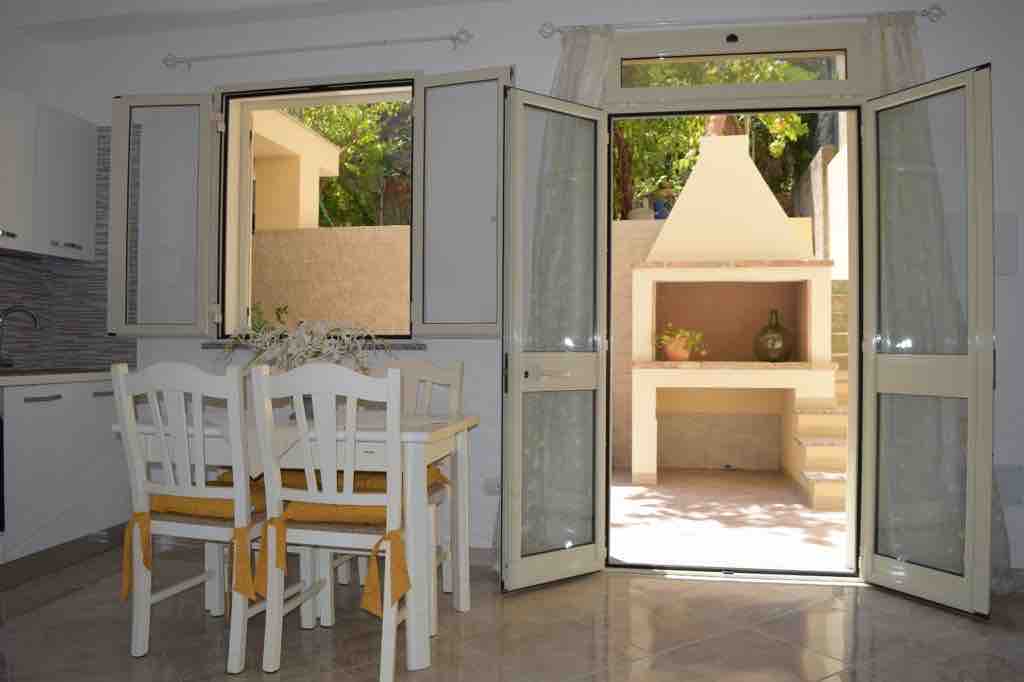 A bright dining area includes a wooden table surrounded by four chairs, with sunlight flooding in from two open doors. A view of a private garden is visible, featuring a stone barbecue and a shaded seating area.
