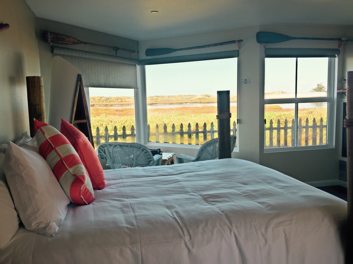 A bright guestroom is featured with a spacious bed dressed in white linens, complemented by vibrant throw pillows. Panoramic windows offer views of the wetlands, while wicker chairs provide a cozy seating area, enhancing the inviting feel of the space.