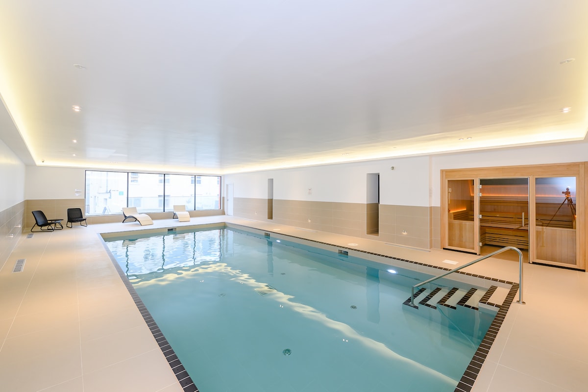 An indoor pool is showcased, featuring a calm aqua surface reflecting soft lighting from the ceiling. Lounge chairs are positioned along the pool's edge, while a sauna is visible in the corner, designed for relaxation.