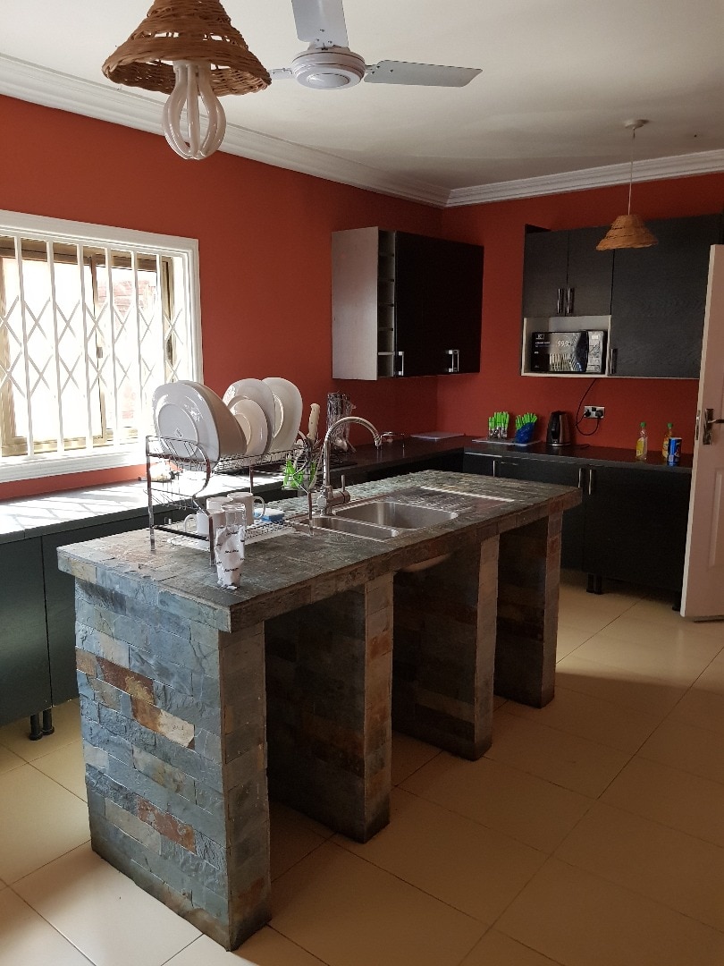 The kitchen features a stone island with a double sink and is complemented by dark cabinetry. A window allows natural light, while dishes are neatly arranged on a drying rack. The walls are painted in a warm tone, and light fixtures hang above.