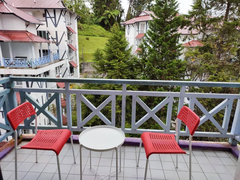 A balcony is shown, featuring two red chairs and a small round table. Lush greenery is visible in the background, providing a sense of tranquility. The railing displays a geometric design that complements the surroundings.