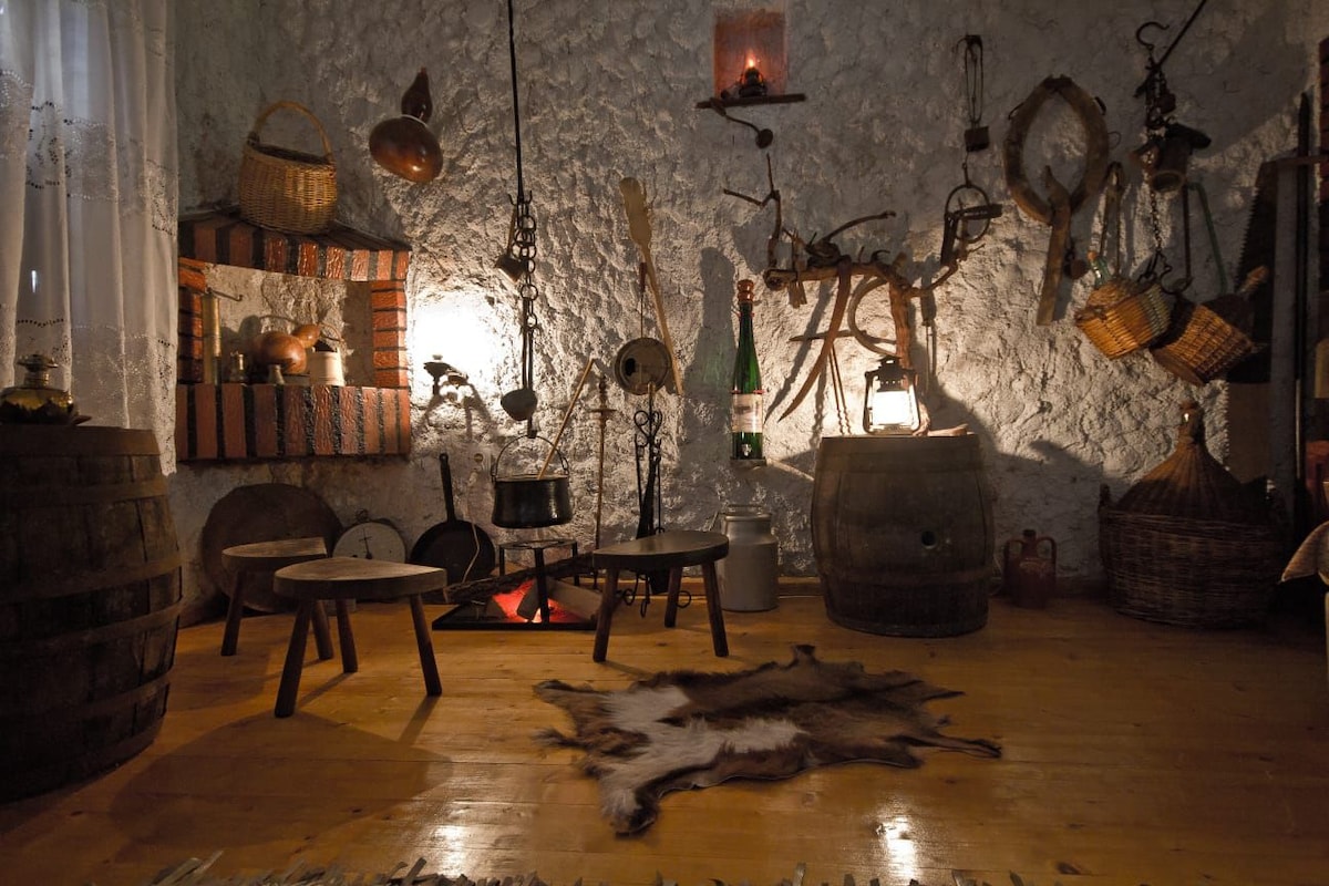 An inviting interior showcases rustic decor, including vintage items displayed on a textured wall. Various containers and tools are arranged on shelves and hung from the ceiling. A warm glow emanates from lanterns, while a soft animal hide is laid out on the wooden floor.