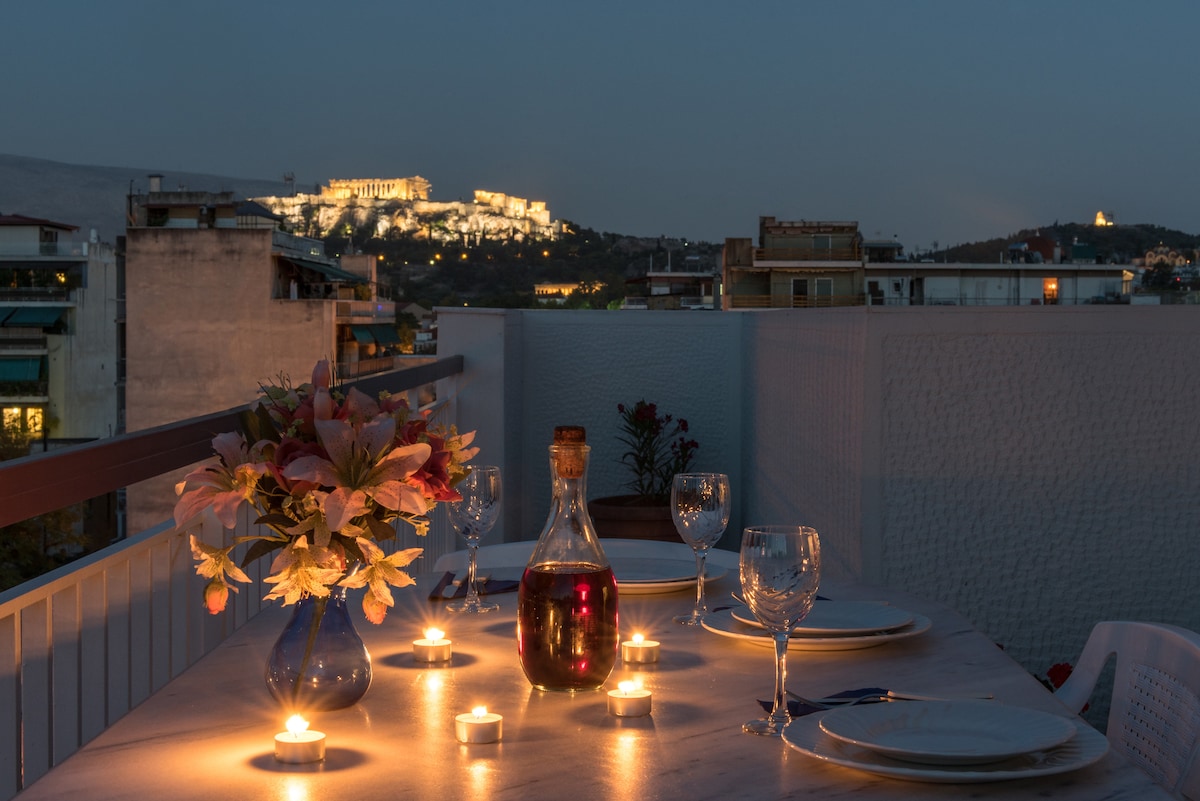 A dining setup is presented on a terrace with a view of illuminated historic buildings in the distance. A marble table is adorned with a vase of flowers, wine bottle, and candles casting soft light, creating an inviting atmosphere for meals outdoors.