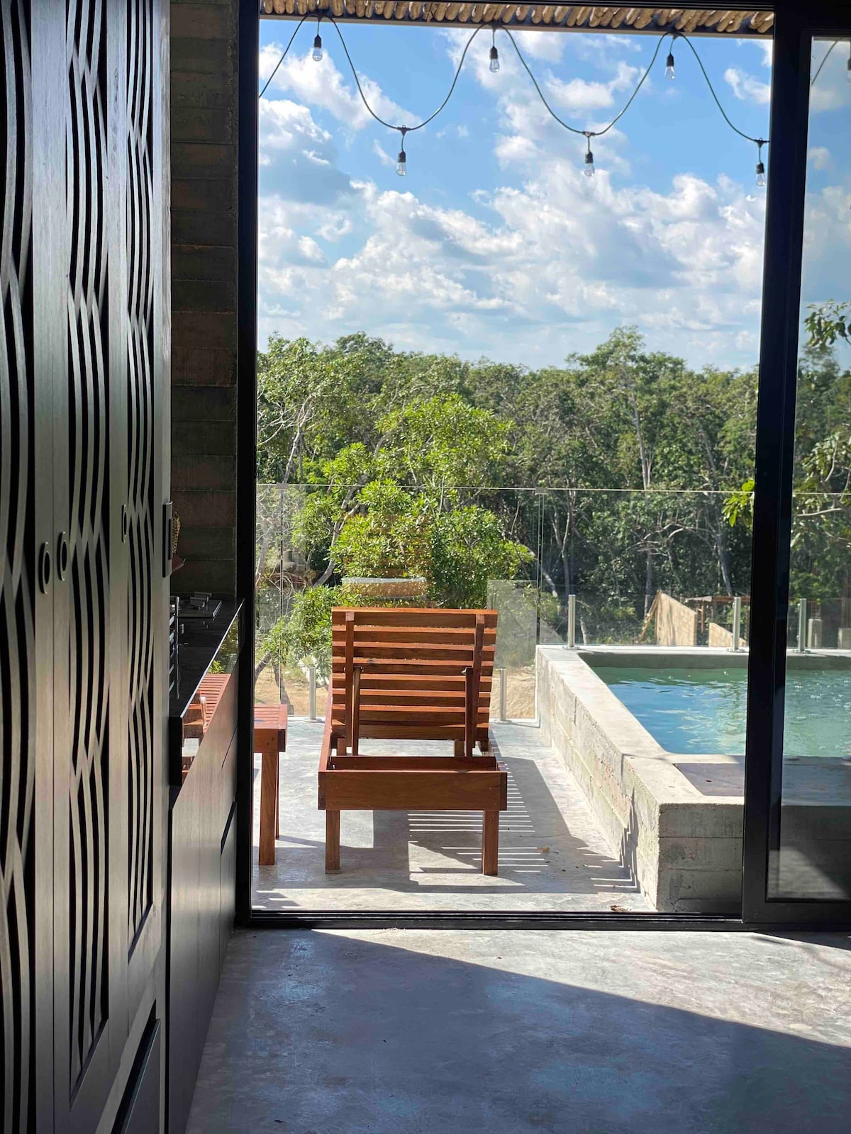 A wooden lounge chair is positioned on a balcony, with views of lush greenery seen through expansive glass doors. The private plunge pool is visible alongside the chair, set against a backdrop of bright blue skies and scattered clouds.