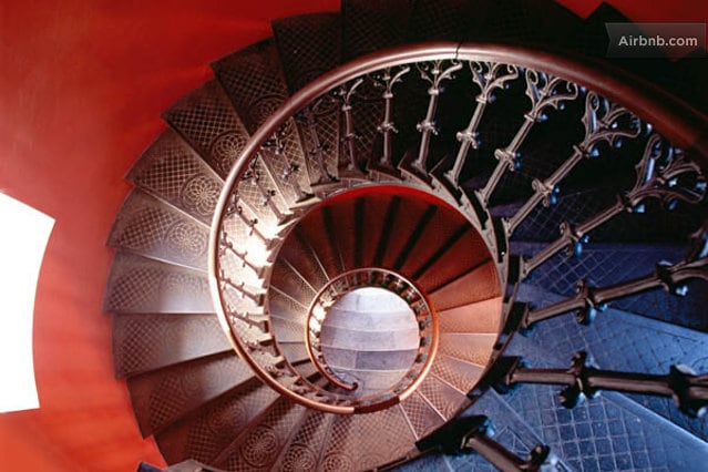 A spiral staircase featuring intricate wrought iron railings and a contrasting color scheme. The stairs appear to descend into a warmly lit area, with a soft glow visible from the top, enhancing the architectural details of the staircase.