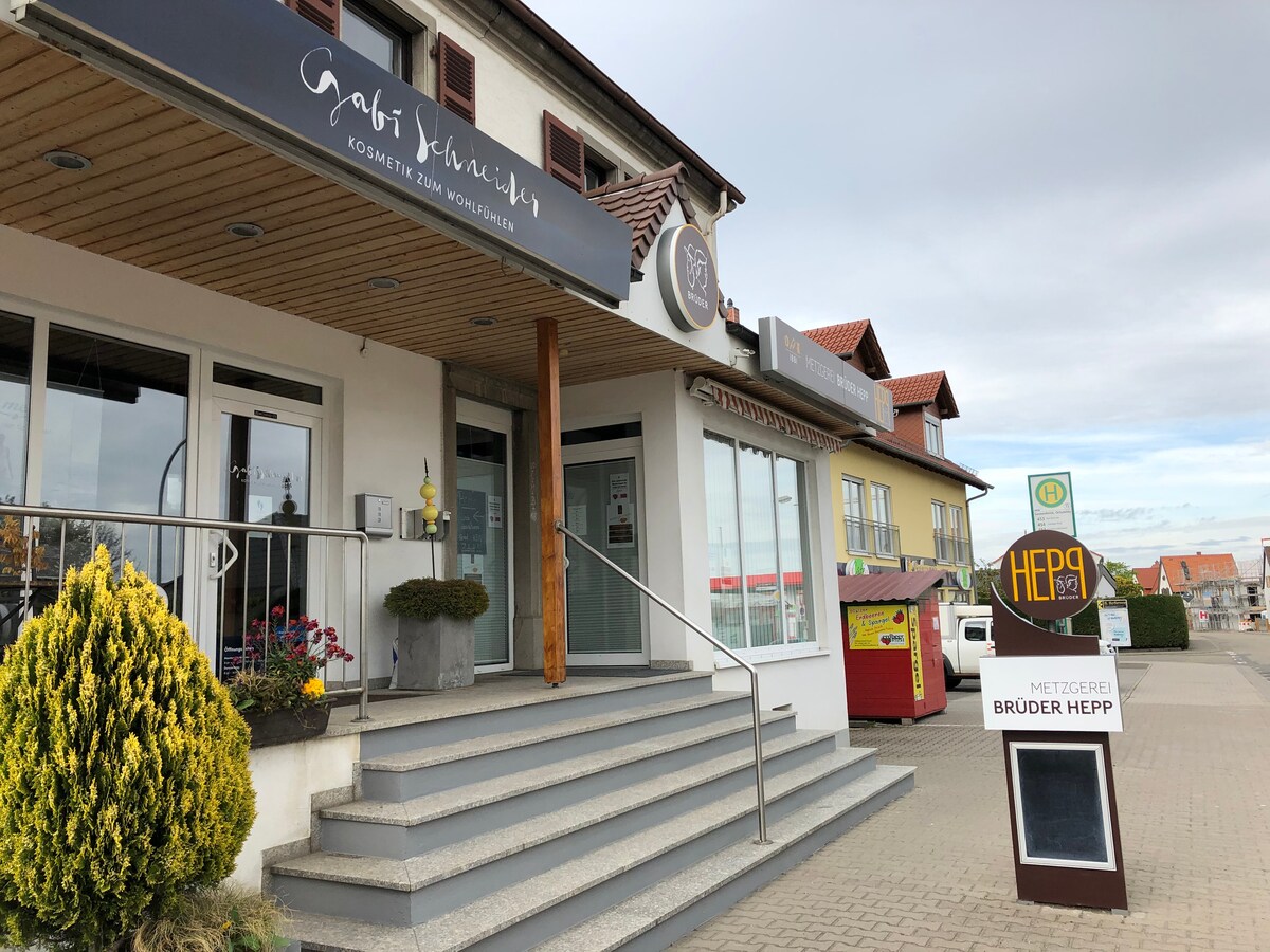 The exterior of a multi-story building is shown, featuring a sloped roof and large glass entrance doors. A sign for a cosmetic studio is visible above the entrance, while a butcher shop is located to the right. Steps lead up to the main door.
