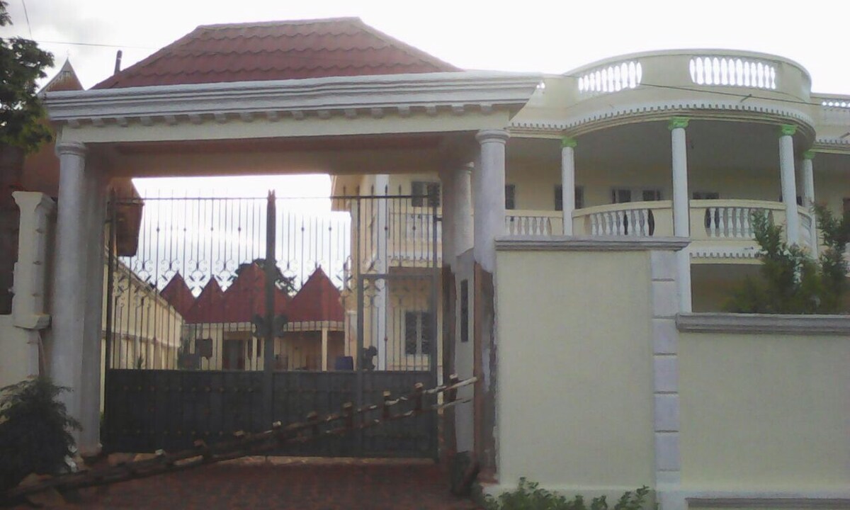 A gated entrance is featured, showcasing a spacious driveway lined with manicured greenery. The gate is flanked by tall columns, leading to a grand architectural structure with a distinct red tiled roof and multiple balconies, enhancing the entrance's impressive appearance.