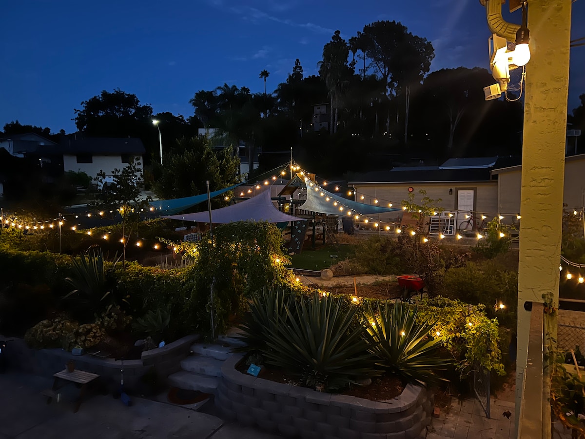 A serene outdoor space is depicted, featuring a canopy of blue fabric and warm white string lights illuminating the area. Lush greenery, including various plants, can be seen, alongside a well-maintained garden backdrop. The peaceful evening sky casts a soft glow over the setting.