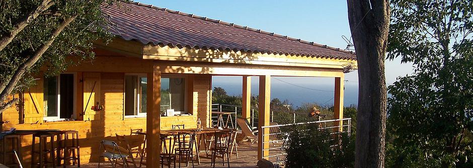 A modern wooden house is featured, surrounded by trees and a spacious deck. Multiple outdoor tables and chairs are positioned on the terrace, allowing for dining and relaxation. The expansive views of the sea are visible beyond the house, highlighting its tranquil setting.