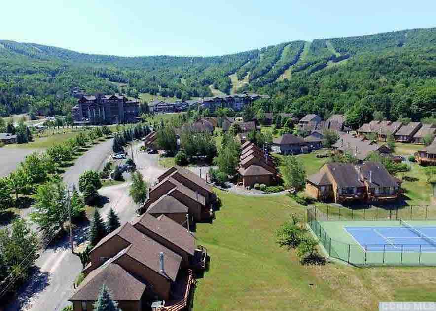 An aerial view captures the layout of the Windham Mountain Village, showcasing rows of cozy townhouses surrounded by lush greenery. In the background, the slopes of the mountain are visible, along with nearby tennis courts and the main lodge, emphasizing the serene yet active atmosphere of the location.