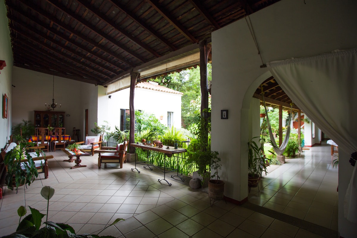 A spacious communal area features a high wooden ceiling and an open layout. Natural light illuminates plants placed throughout. Comfortable seating arrangements with wooden furniture invite relaxation, while a small dining table is visible. Arched doorways lead to the garden, enhancing the tranquil atmosphere.