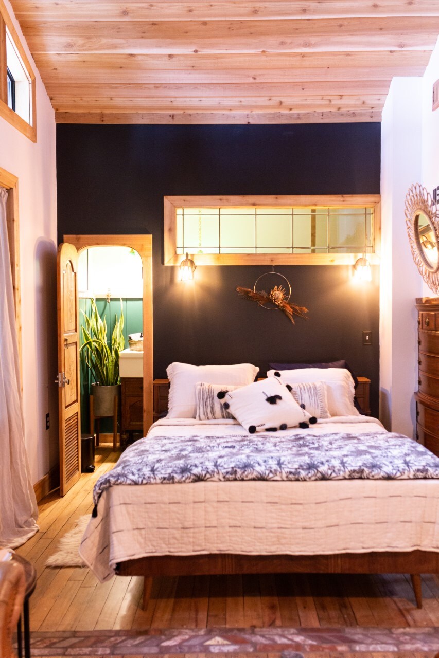 The cozy sleeping area features a spacious bed adorned with a textured white blanket and patterned pillows. Natural wood paneling complements the high ceiling, while a wooden dresser provides storage. A subtle plant and a mirror enhance the calming ambiance of the room.