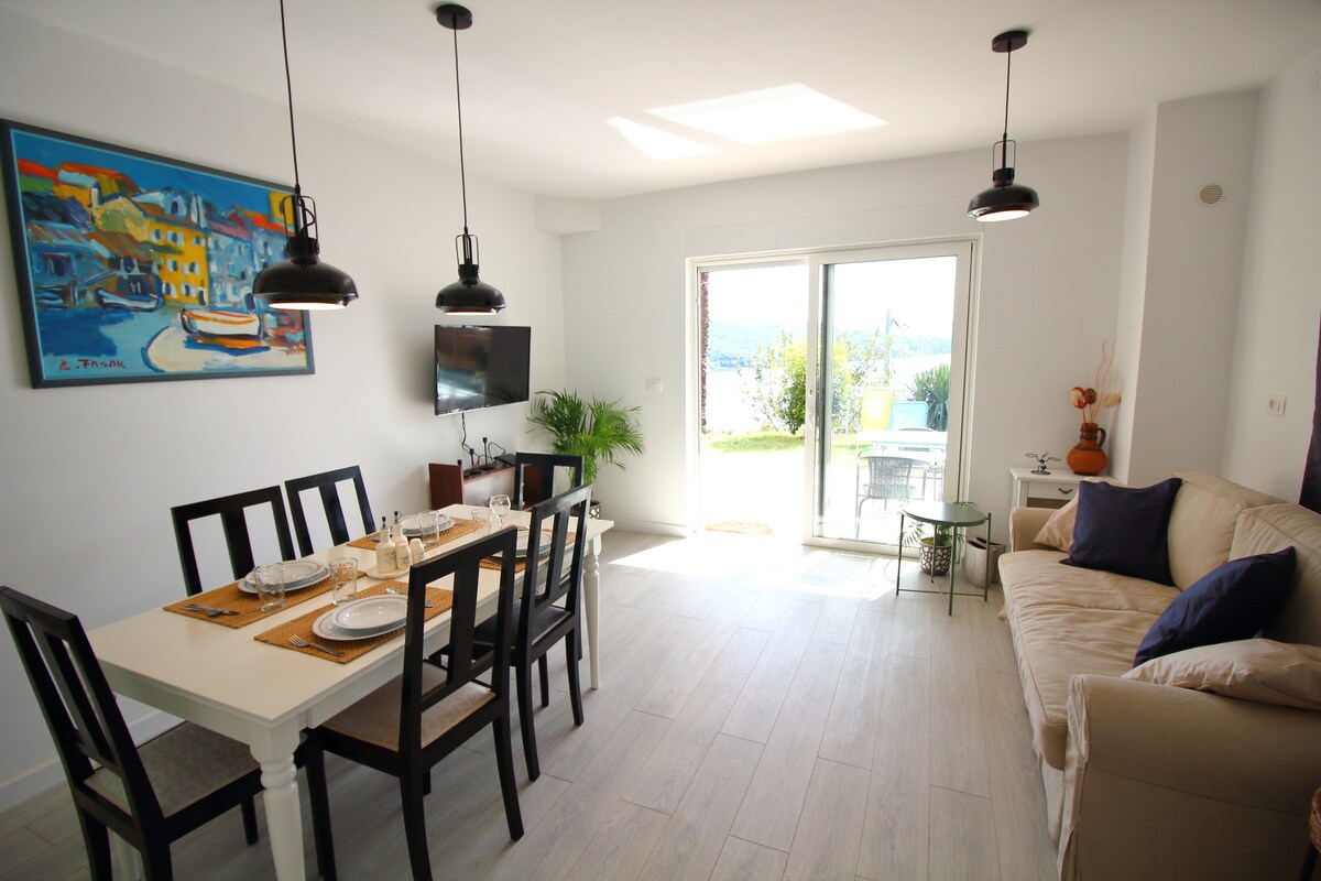A light-filled dining area features a long table set for six, accompanied by black chairs. A cozy sofa stands nearby, and a flat-screen television is mounted on the wall. Large sliding doors open to the outside terrace, connecting the interior to the garden.