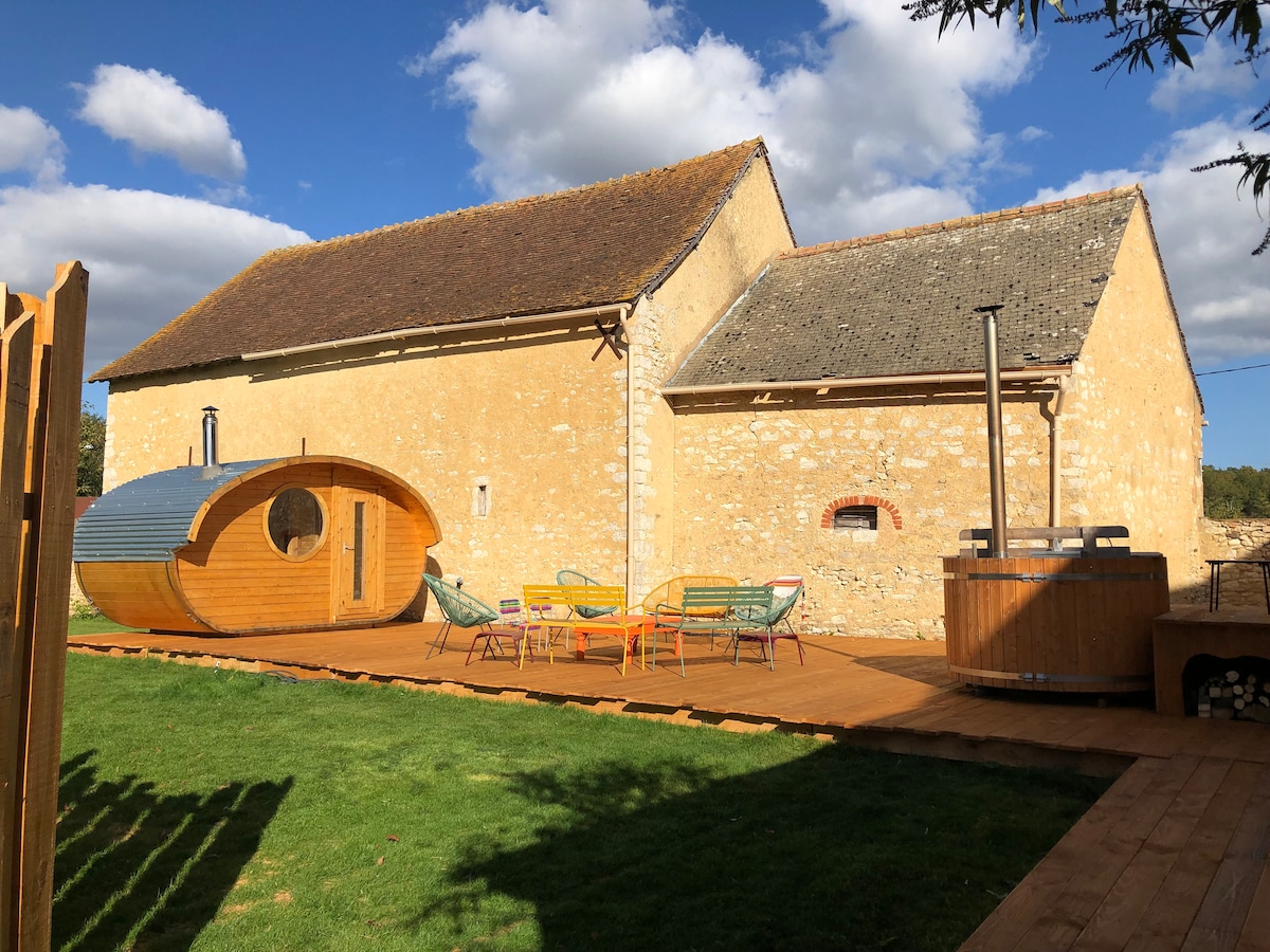 The exterior of a rustic stone building is complemented by a wooden sauna pod and hot tub, set on a spacious wooden deck. Colorful lounge chairs surround a central table, with a grassy area providing a tranquil setting. The sky features scattered clouds.