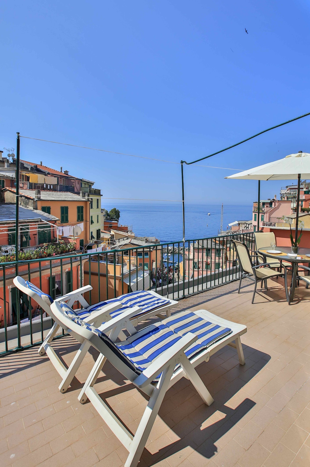 A spacious terrace offers comfortable seating with striped lounge chairs and a dining area. The sea view is unobstructed, showcasing the calming blue water and colorful buildings of Riomaggiore. An umbrella provides shade, adding to the inviting atmosphere of the outdoor space.