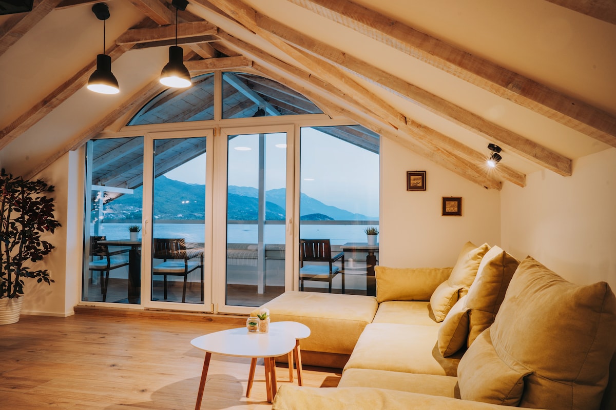 A bright living area features a spacious yellow sectional sofa, offering comfort and warmth. Large glass doors reveal a scenic view of Ohrid Lake and the mountains beyond, while exposed wooden beams add character to the ceiling.