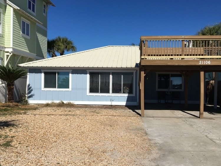A charming blue exterior of the house is visible, featuring large windows that allow natural light to enter. A spacious deck above offers outdoor seating. The driveway is lined with gravel and is accompanied by tropical landscaping, creating a welcoming beach home appearance.