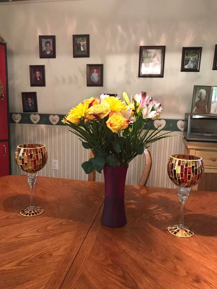 A dining table is set with a vibrant bouquet of yellow and pink flowers in a purple vase, positioned at the center. Two decorative goblets with mosaic designs are placed on either side of the arrangement. Family photos adorn the walls in the background.