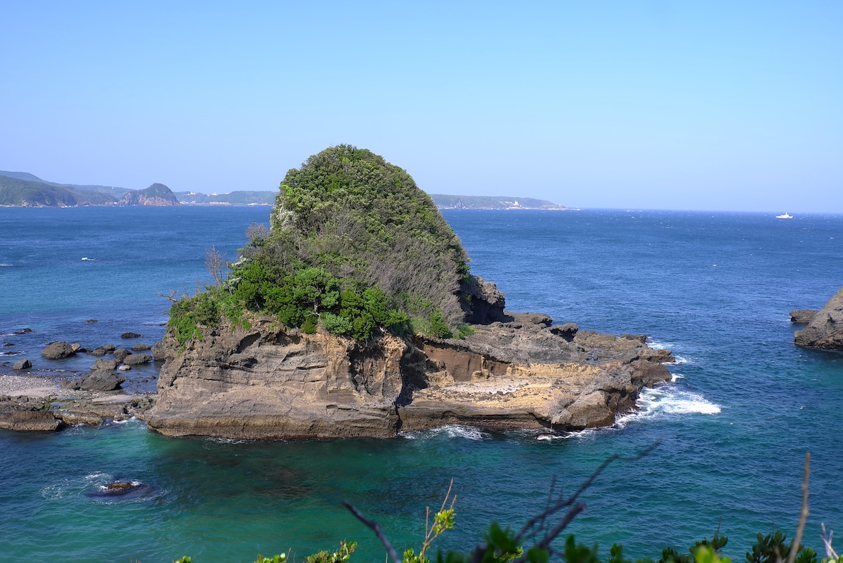 A rocky island emerges from turquoise waters, covered with lush green vegetation. Gentle waves lap against the shoreline, showcasing a serene coastal landscape. In the background, distant hills and a clear blue sky create a peaceful ambiance.