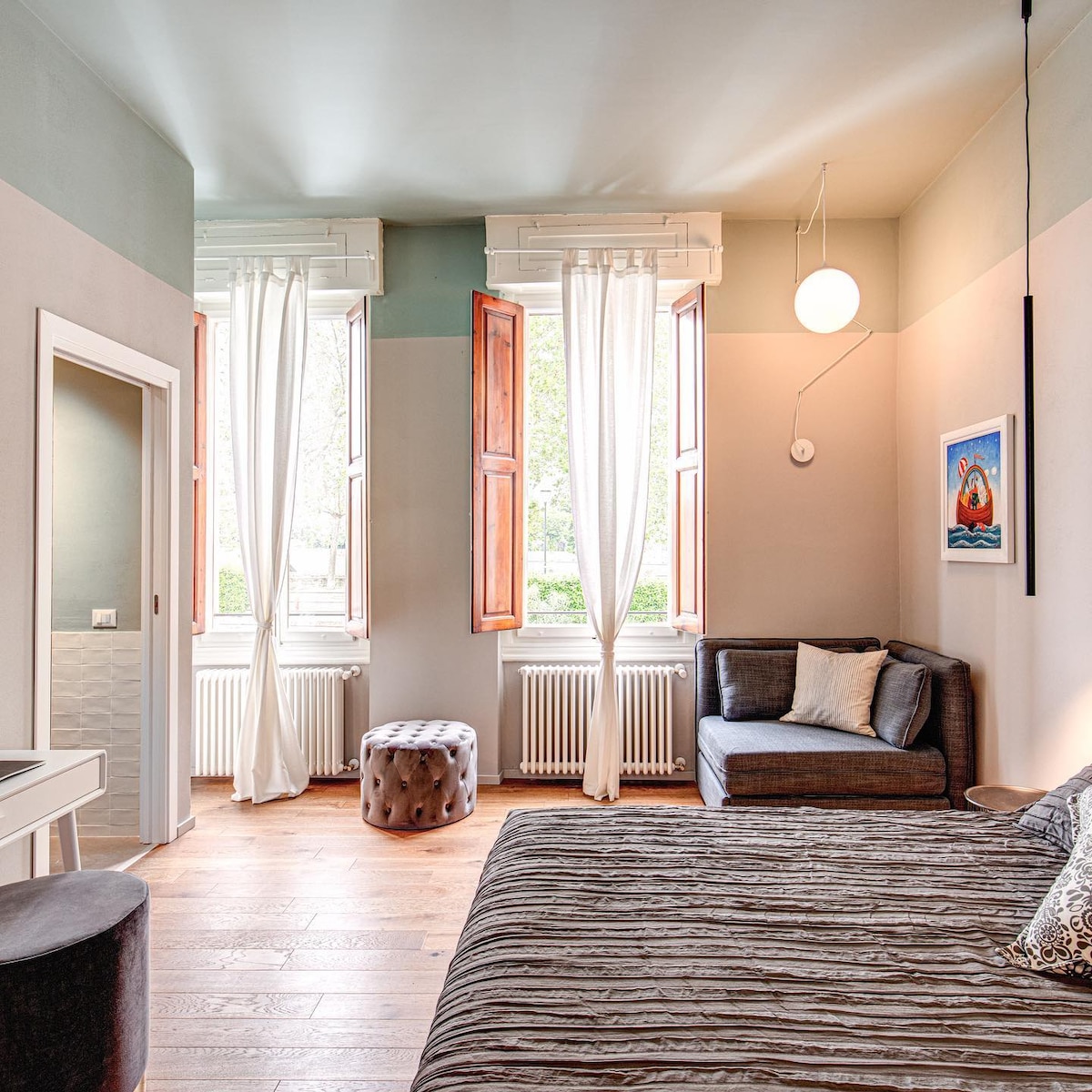 A well-appointed room features large windows with wooden frames, allowing natural light to fill the space. A spacious bed is complemented by cozy seating in one corner. Light-colored walls enhance the airy atmosphere, while a piece of artwork adds a touch of color.