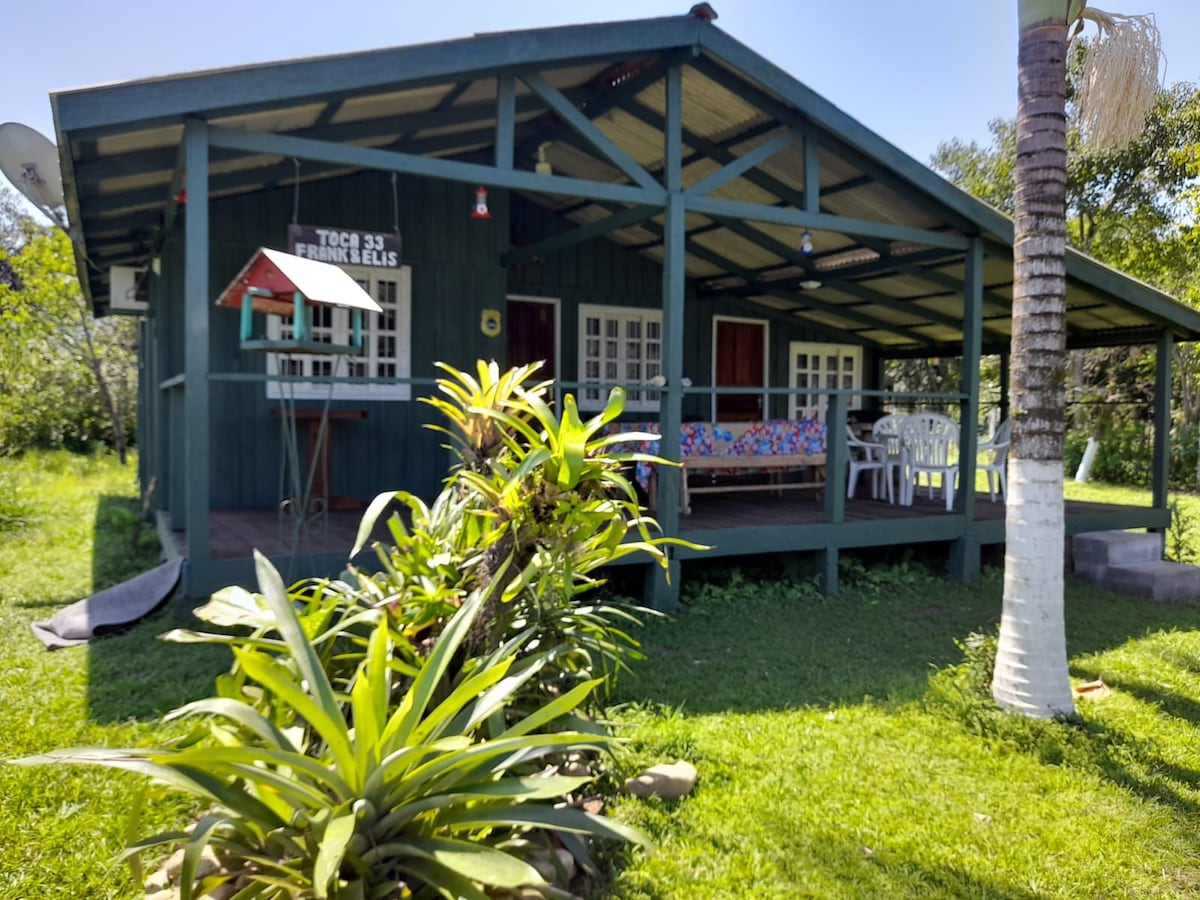 A charming wooden cabin features a large covered porch, accented by a wooden bench and colorful seating. Lush green grass surrounds the house, with tropical plants and trees enhancing the natural setting. Large windows allow ample light inside, creating a bright and open environment.