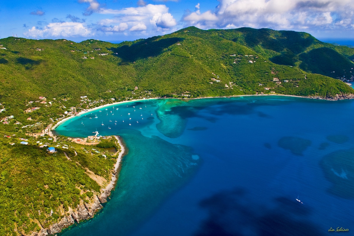 Aerial view of a picturesque bay surrounded by lush green hills. Clear turquoise waters gently lap against sandy shores, with white sailboats scattered across the sea. The coastline features a blend of sandy beaches and rocky outcrops, enhancing the natural beauty of the landscape.