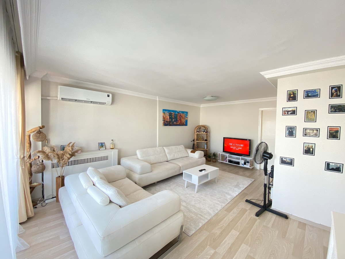 A bright living area is filled with natural light from large windows. Comfortable, white sofas are arranged around a low coffee table. A mounted television and decorative shelves add to the space, while framed photographs create visual interest on the nearby wall.