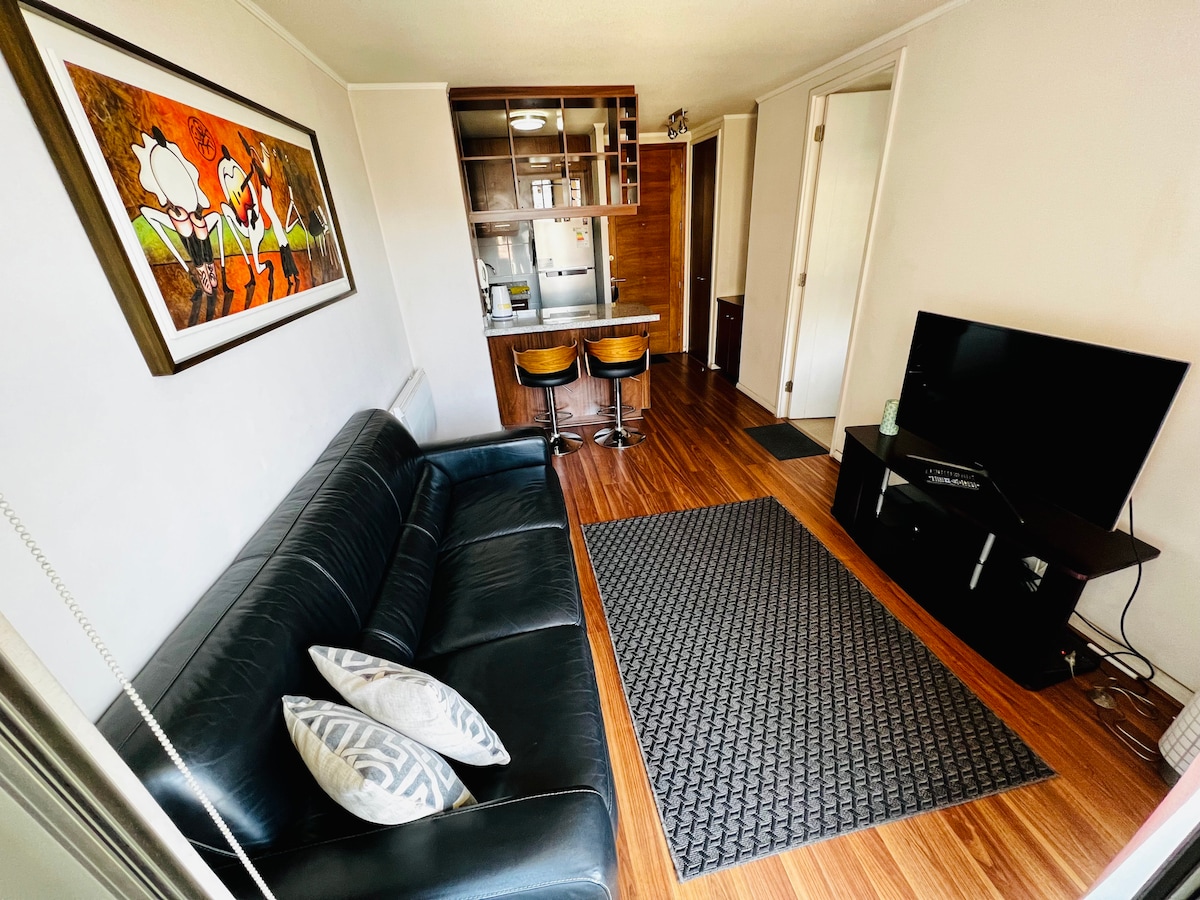 A living area is presented with a modern black sofa featuring decorative pillows. A wall art piece adds a touch of creativity above a wooden dining table with two stools. A television is visible on a low stand, and the space is illuminated by natural light.