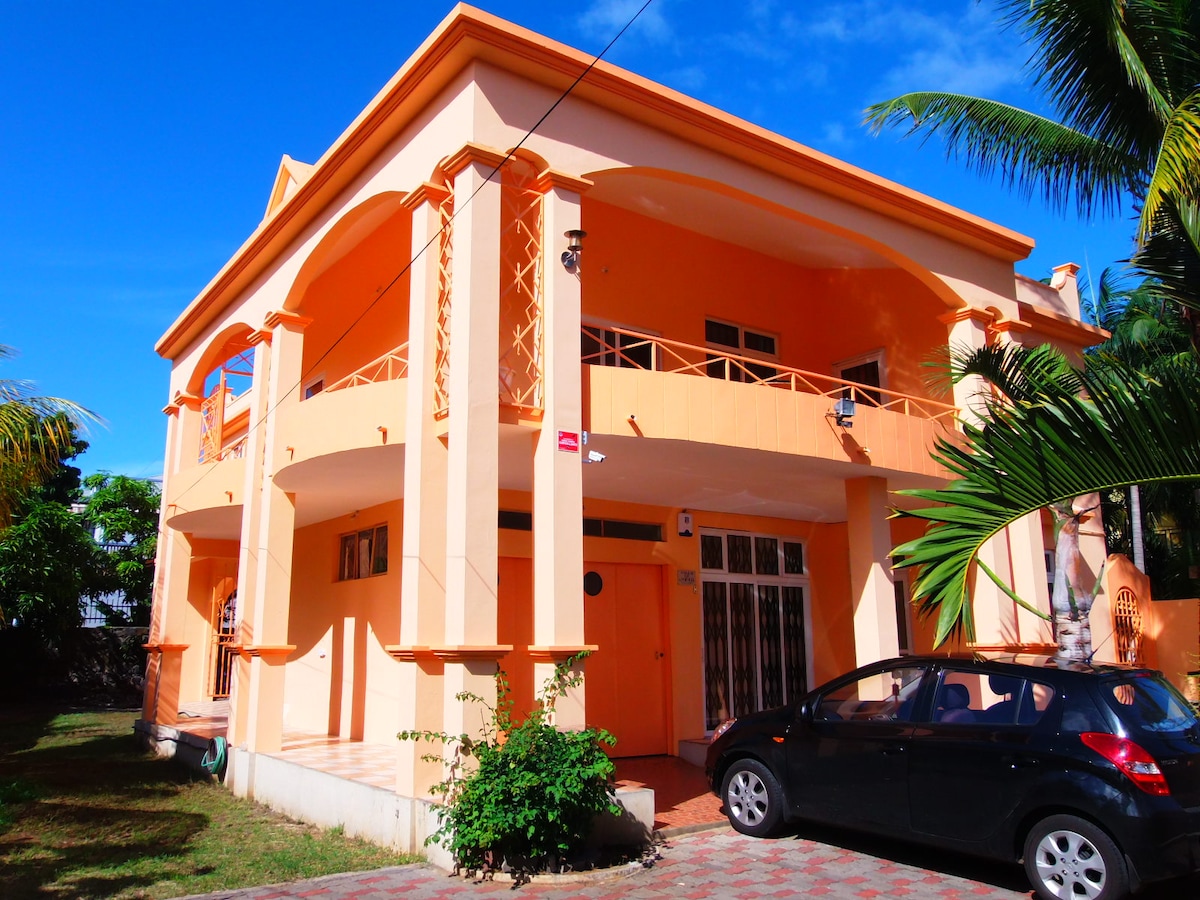 A vibrant two-story house features an orange exterior with archways and a spacious front porch. A black car is parked in front, and lush greenery surrounds the property, contributing to a welcoming atmosphere. The clear blue sky complements the bright façade.