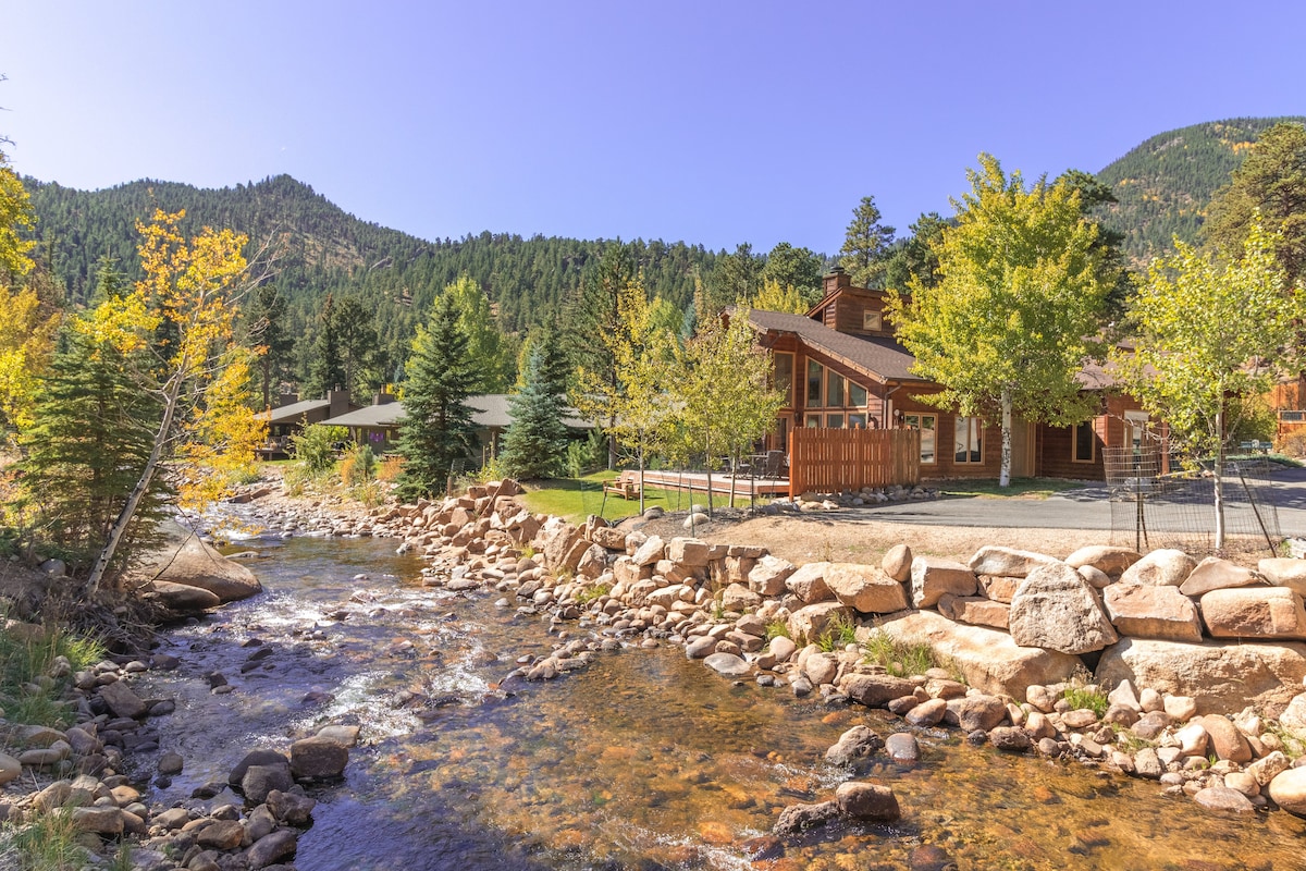 A picturesque riverside view showcases a well-maintained property with rustic wooden architecture. The gently flowing water reflects the surrounding greenery, while large rocks line the riverbank. Mountainous terrain rises in the background under a clear blue sky, highlighting the tranquil setting.