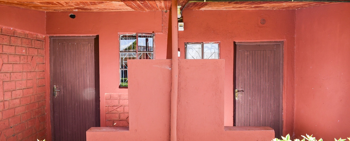The exterior of a red brick building is shown, featuring two separate entrances. Each door is framed by a simple wooden structure. Small windows above each door allow natural light to illuminate the space. The surrounding area is neatly maintained with foliage visible at the base.