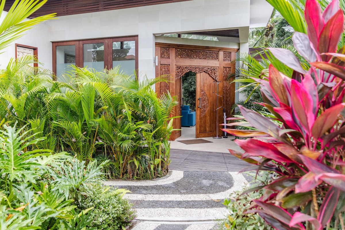 An entrance is framed by lush tropical foliage, featuring vibrant green and reddish leaves. The pathway, made of stone, leads towards intricately carved wooden doors, creating an inviting entryway to the villa.