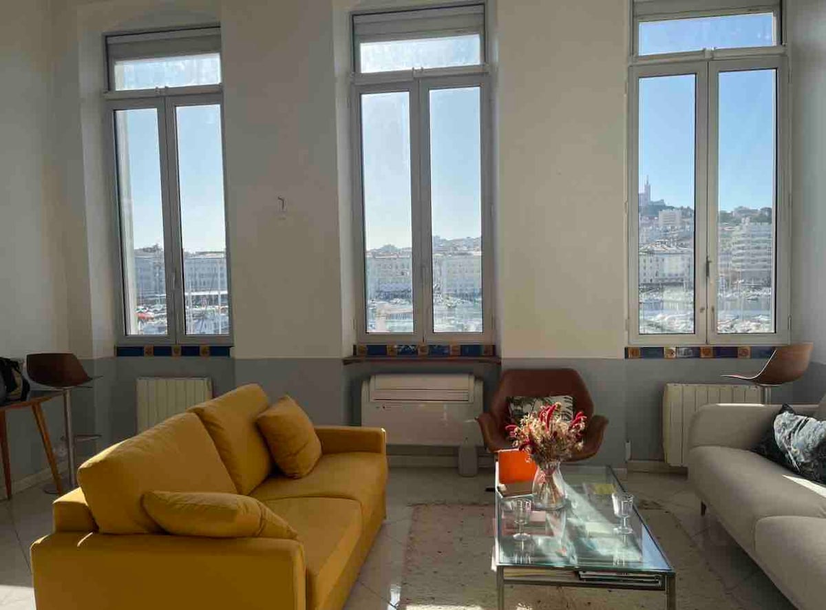 Large windows frame a view of the surroundings, allowing natural light to fill the space. A bright yellow sofa contrasts against neutral walls, paired with a glass coffee table holding a floral arrangement. Two modern chairs complement the seating area.