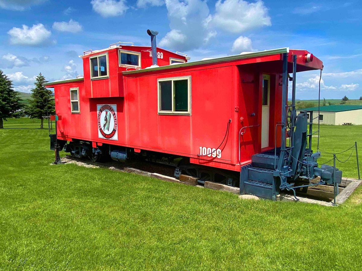 The exterior of a bright-red Northern Pacific caboose is displayed on a grassy area, showcasing its original features. The structure includes a cupola, multiple windows, and vintage railroad details, all set against a clear blue sky with fluffy clouds.