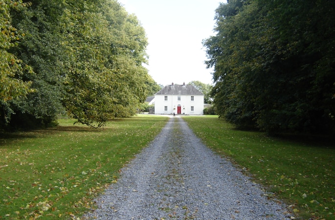 A gravel drive lined with tall trees leads to an elegant white house set on a manicured lawn. The house features a red door, surrounded by a serene landscape of greenery and scattered leaves, creating a tranquil approach.