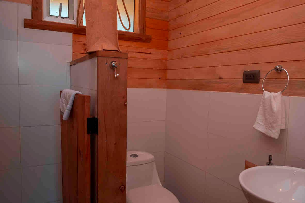 A compact bathroom features wooden accents alongside tiled walls. A modern toilet and a small sink with a towel holder are visible. Natural light illuminates the space through a small window, enhancing the inviting atmosphere.
