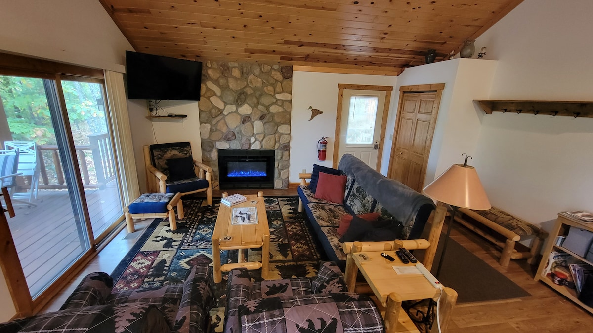 The living area features a rustic design with a stone fireplace and a flat-screen TV mounted on the wall. Comfortable seating includes a full-size couch and two armchairs. Natural light floods in through sliding doors leading to a large deck, enhancing the welcoming atmosphere.